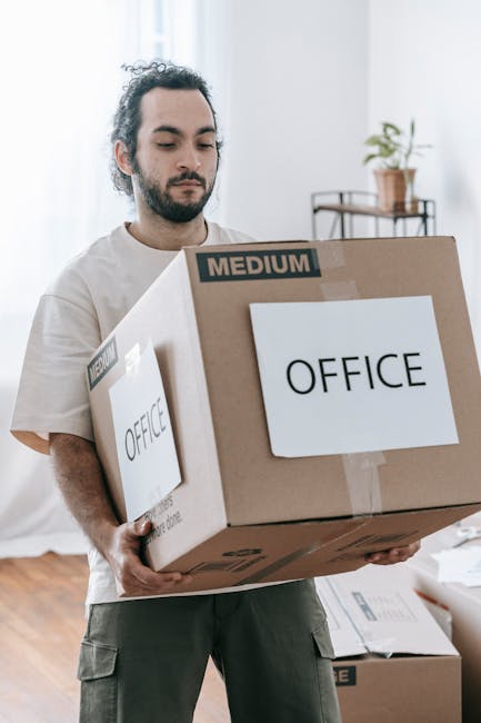A man with dark, curly hair and a beard, dressed in a light-colored t-shirt, is holding a large cardboard box labeled 'OFFICE' in black text with a 'MEDIUM' size indicator on it. He is inside a bright