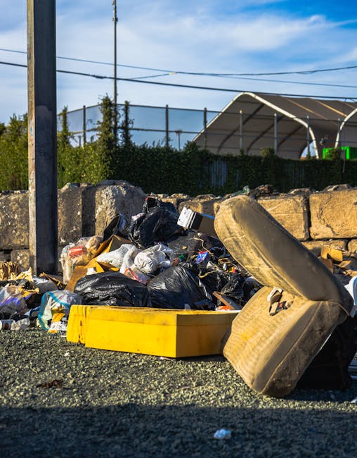 Close-up view of discarded rubbish behind a black metal fence, featuring crumpled plastic bags, paper packaging, cardboard, and other household waste materials scattered on the ground amidst dry leave