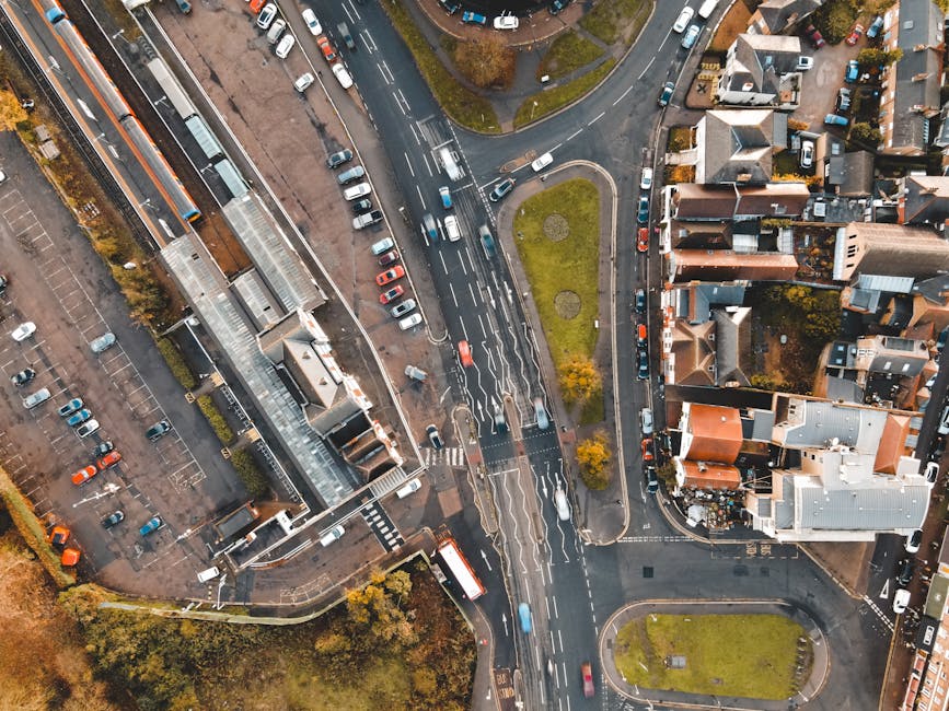 An aerial view of Raynes Park station area showing multiple lanes of traffic on the road, with parked cars along the streets and in a large parking lot adjacent to the station building. The station fe