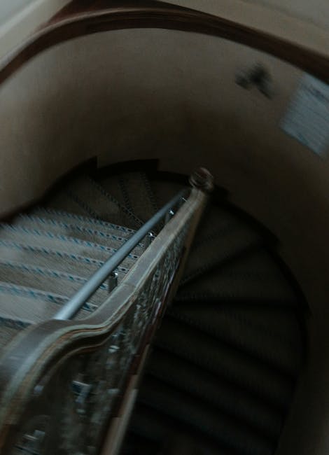A view of a narrow staircase inside a residential property with green carpeted stairs and wooden handrails on both sides, supported by turned wooden balusters. The staircase curves downward out of sig